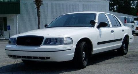 2000 Ford Crown Victoria - A white sedan is parked in front of a building with a palm tree nearby. Another white van is visible in the background.