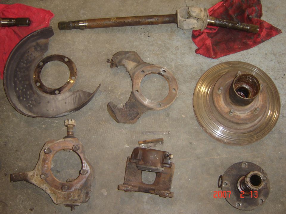 Disassembled front suspension parts laid out on concrete, including a brake rotor, spindle, caliper, and CV axle