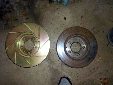 1997 Lincoln Town Car - A new, gold-colored slotted brake rotor is shown next to a used, rusted brake rotor on a concrete surface.
