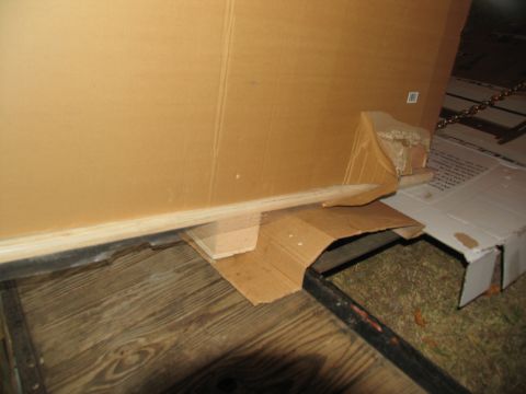 1988 Ford C-350 (Centurion Conversions) - A close-up view of a damaged cardboard box resting on a wooden floor, with a metal frame visible below.