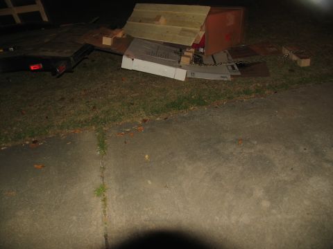 1988 Ford C-350 (Centurion Conversions) - A trailer sits on grass at night, covered in various discarded materials and a makeshift wooden structure.