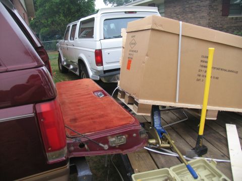 1988 Ford C-350 (Centurion Conversions) - A maroon truck's tailgate is open, revealing its carpeted interior, with a white SUV and a large boxed item on a pallet nearby.
