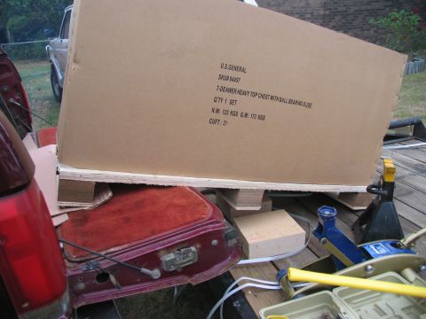 1988 Ford C-350 (Centurion Conversions) - A large cardboard box labeled U.S. GENERAL is loaded onto a trailer, with parts of a red vehicle visible.