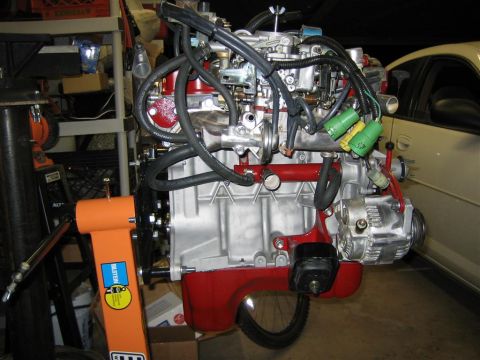 1987 Suzuki Samurai - Engine assembly mounted on an orange stand, with visible hoses, wiring, and a portion of a car in the background.