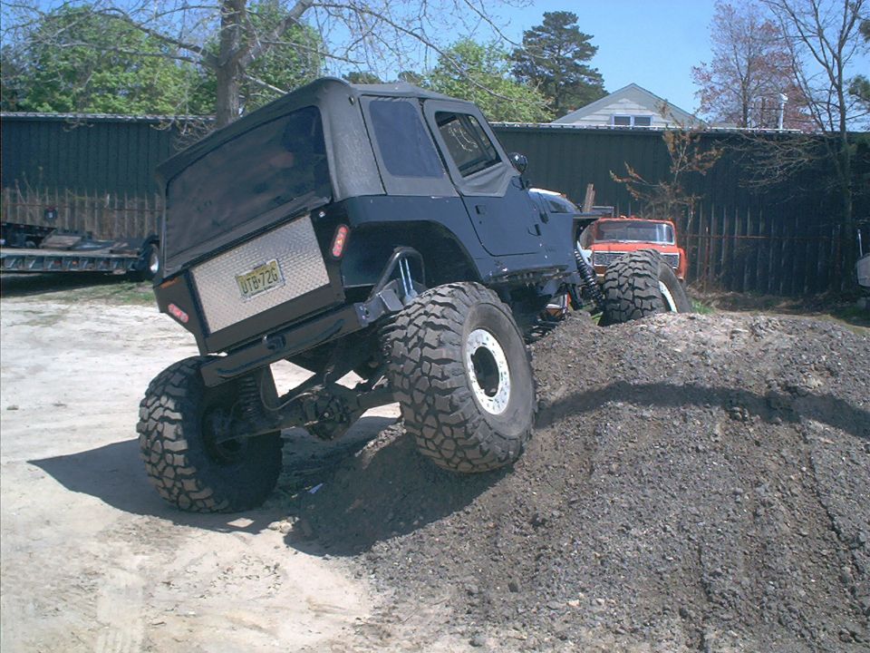 Black Jeep Wrangler with large tires climbing a dirt mound, diamond plate storage box on rear