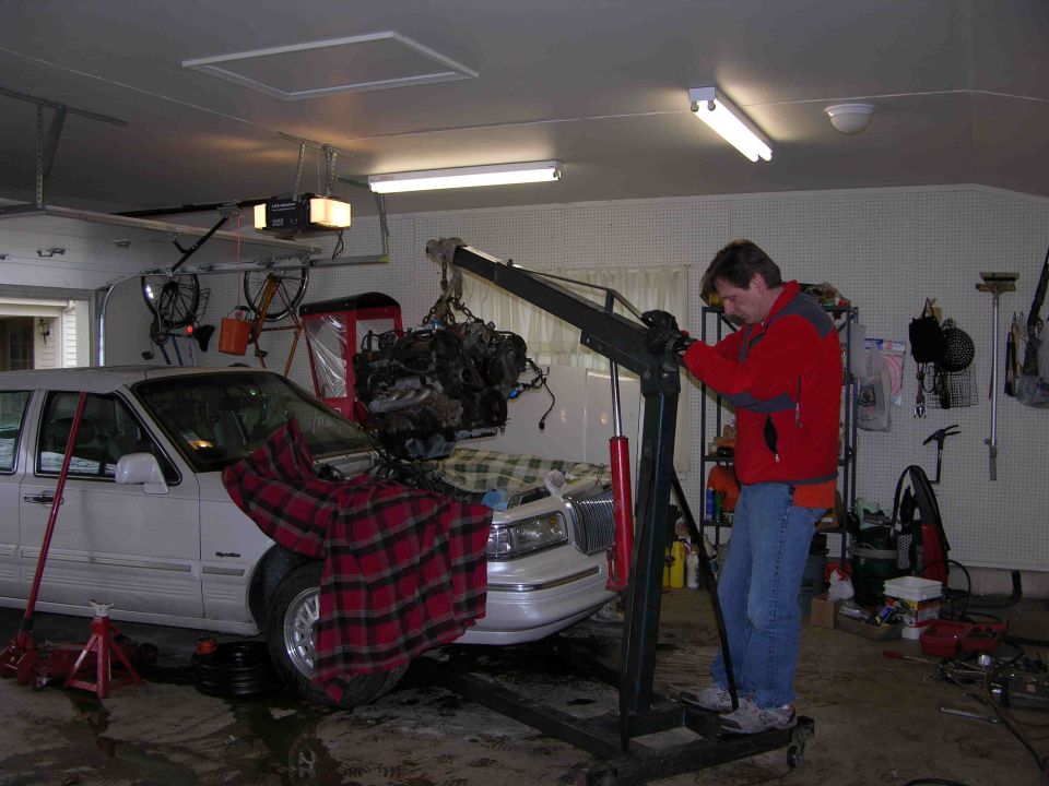 Man using engine hoist to pull a V8 from a white sedan in a garage