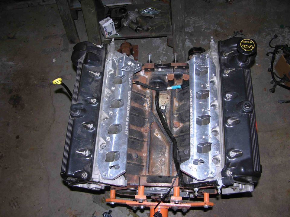V8 engine on orange stand with black valve covers, intake removed, in a garage.