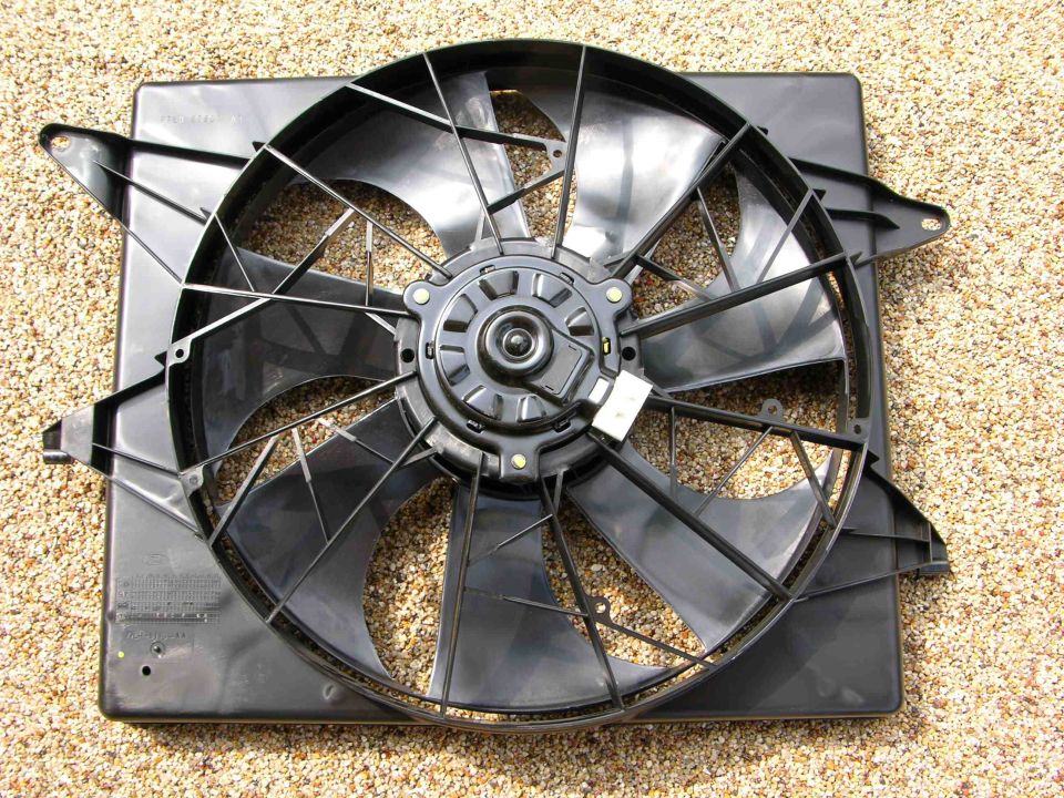Car radiator fan assembly with black plastic blades and a central motor housing, resting on a gravel surface