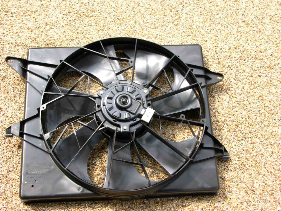 Black plastic radiator fan assembly with a central motor housing, lying on a gravel surface