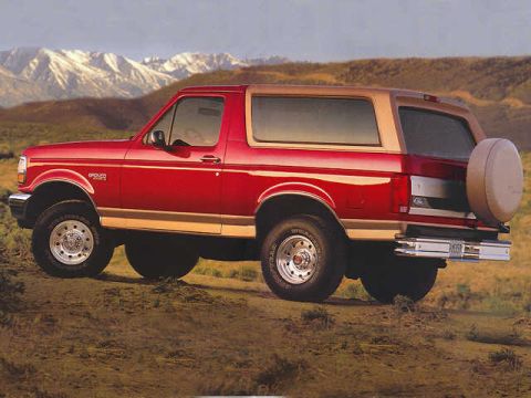 1994 Ford Bronco - A red 1994 Ford Bronco with a tan top is shown from a three-quarter front angle, parked in a dry, mountainous landscape.