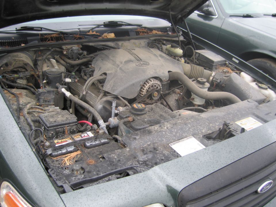engine bay of a Ford Crown Victoria, hood up, showing engine and battery. Another Crown Victoria is parked next to it