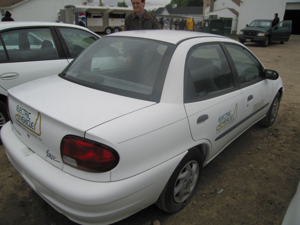 White electric sedan with 'Electric Vehicle' decals on the side and rear. Another white car is parked next to it