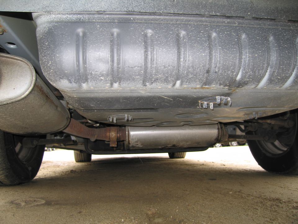 Underside of a vehicle showing the exhaust system and fuel tank. The exhaust pipe is rusty