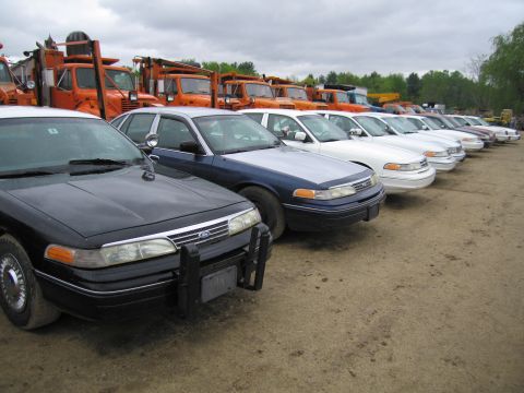 1991 Ford LTD Crown Victoria - Several police cars are parked in a lot, with orange trucks in the background.