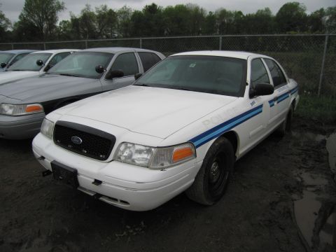 1991 Ford LTD Crown Victoria - A white police car with blue stripes is parked on muddy ground next to other vehicles.