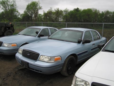 1991 Ford LTD Crown Victoria - Two light blue police cars are parked side-by-side in an outdoor lot under a cloudy sky.