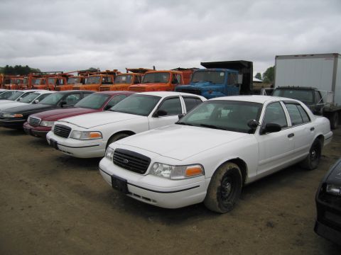 1991 Ford LTD Crown Victoria - Several white Ford Crown Victoria sedans are parked in a lot with various trucks in the background.