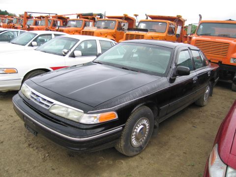 1991 Ford LTD Crown Victoria - A black sedan is parked in a lot, with several orange dump trucks visible in the background.