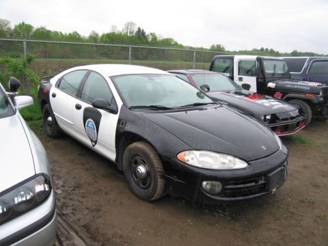 1991 Ford LTD Crown Victoria - A black and white police car is parked on a muddy lot next to other vehicles under a cloudy sky.