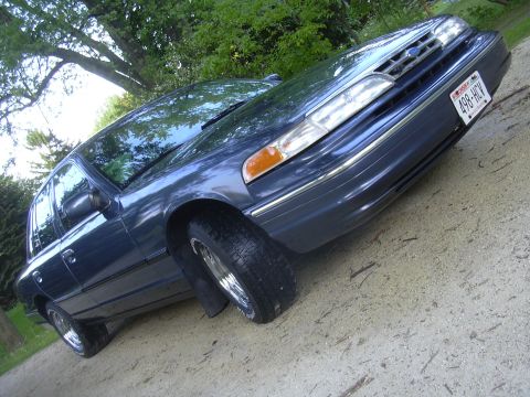 1996 Ford Crown Victoria - A dark blue sedan is parked on a dirt surface with trees in the background.