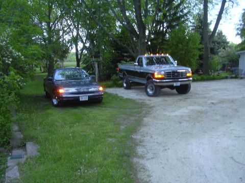 1996 Ford Crown Victoria - A lifted Ford F-250 pickup truck is parked next to a dark colored sedan on a property with trees and a house in the background.