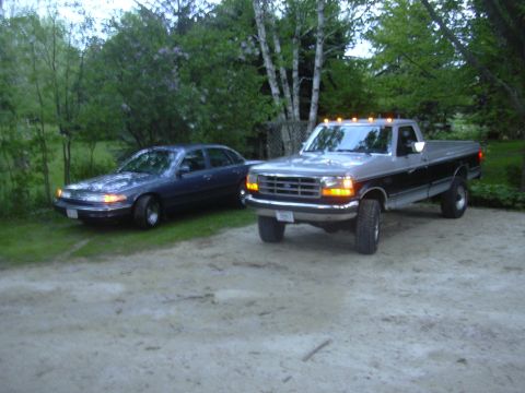 1996 Ford Crown Victoria - Two vehicles are parked on a dirt and grass area with trees in the background.