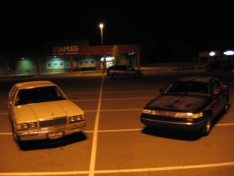 1988 Mercury Grand Marquis - Two sedans, including a 1988 Mercury Grand Marquis, are parked in a lot outside a Staples store at night.