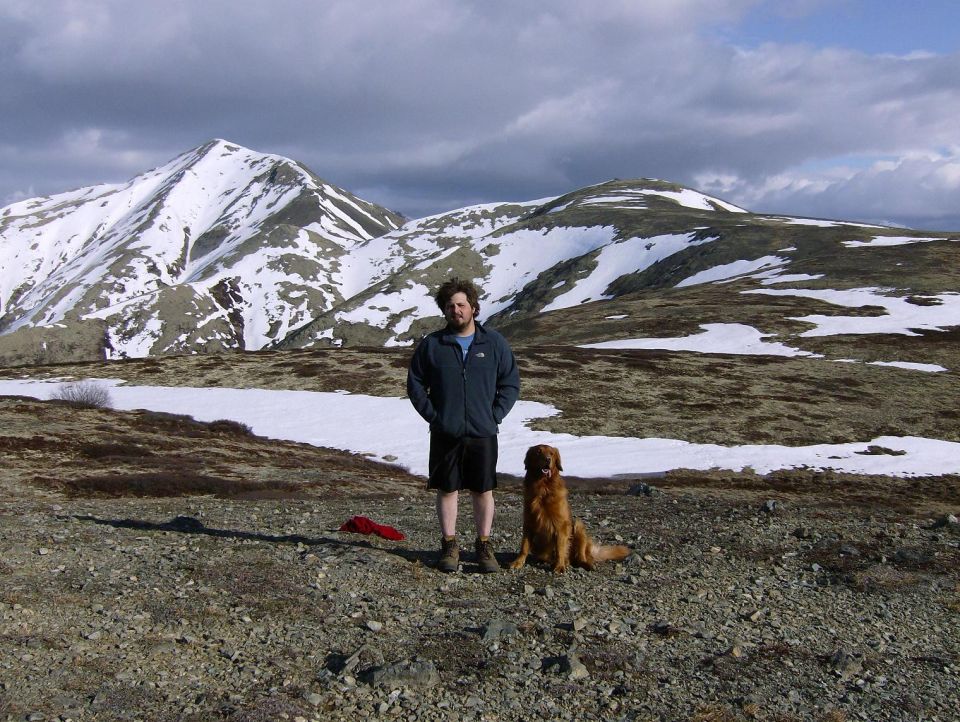 Guy and his dog are standing in a rocky, snowy mountain area. There's a red object on the ground