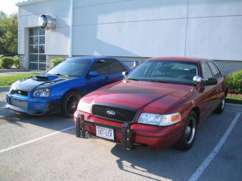 2001 Ford Crown Victoria - A red 2001 Ford Crown Victoria is parked next to a blue Subaru WRX.