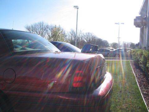 2001 Ford Crown Victoria - The rear of a red sedan is visible, with other vehicles parked in a lot under a bright sky.
