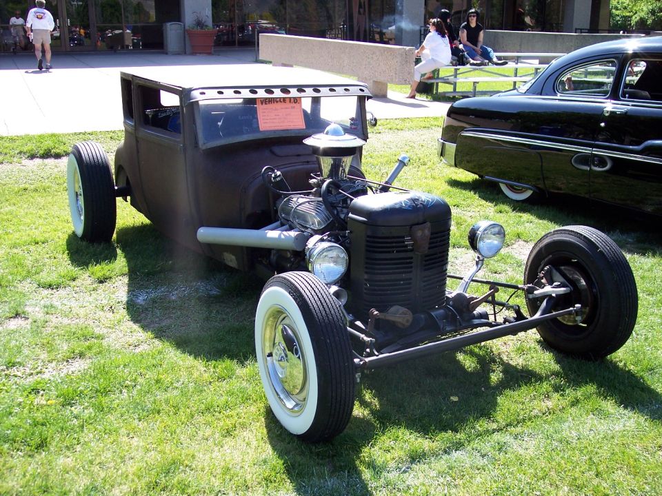 Custom hot rod with exposed engine and whitewall tires, parked on grass next to a black classic car