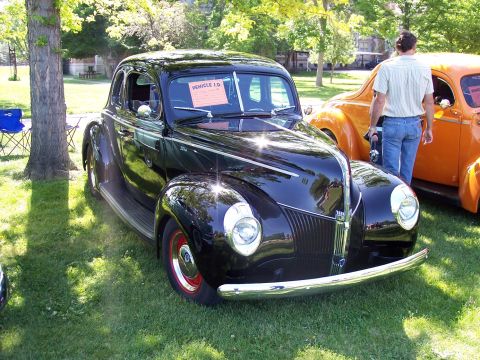 1972 Ford Ranchero - A black coupe with red wheels is displayed at an outdoor car show next to an orange vehicle.