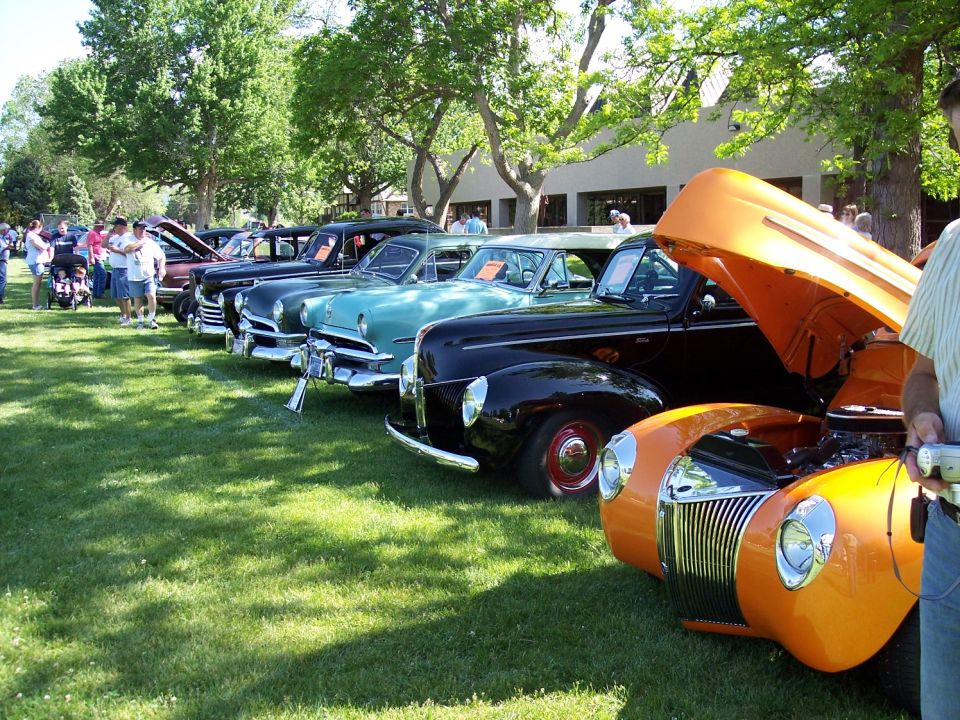 Several classic cars lined up on grass at an outdoor show. Some hoods are open