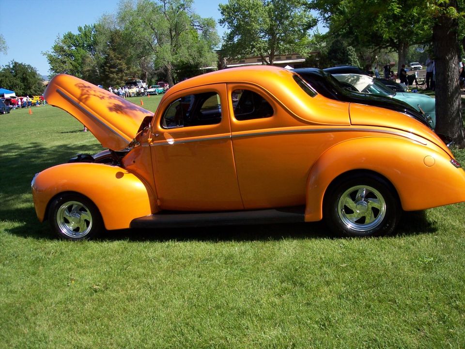 Orange hot rod coupe with hood open, custom flames on hood, chrome wheels, parked on grass at a car show
