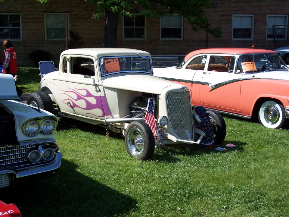 Hot rod coupe with purple flames, exposed engine, and an American flag on the grille. A 1950s sedan is parked behind it