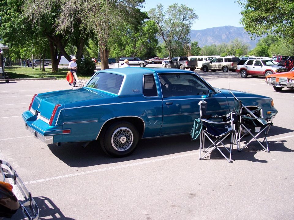 Blue coupe parked in a lot with two folding chairs in front. Other vehicles and mountains in background