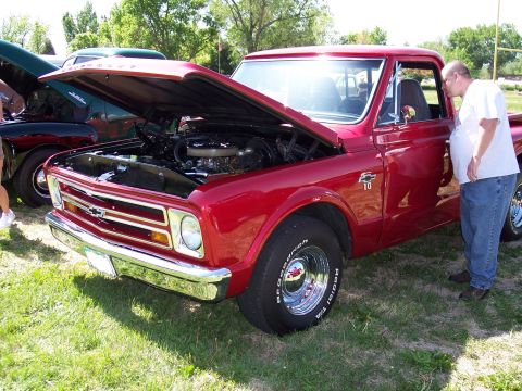 1972 Ford Ranchero - A man inspects a red pickup truck with its hood open at an outdoor car show.