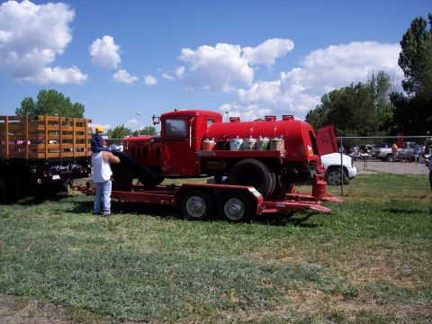 1972 Ford Ranchero - A red tanker truck is being transported on a trailer, with a man and child nearby and other vehicles in the background.