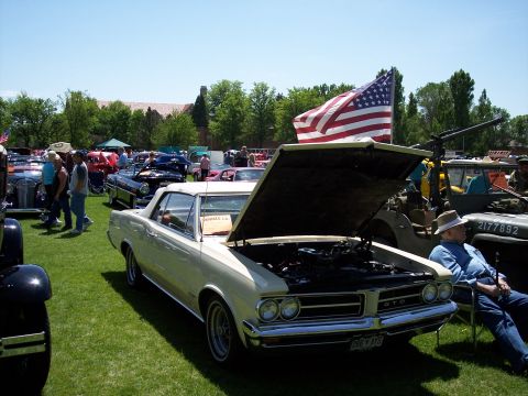 1972 Ford Ranchero - A cream-colored 1965 Pontiac GTO convertible is displayed with its hood open, under a waving American flag at a car show.