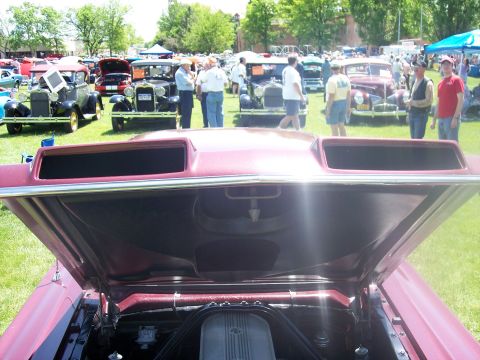 1972 Ford Ranchero - The hood of a red vehicle is open, revealing a prominent hood scoop and engine parts, with a car show in the background.