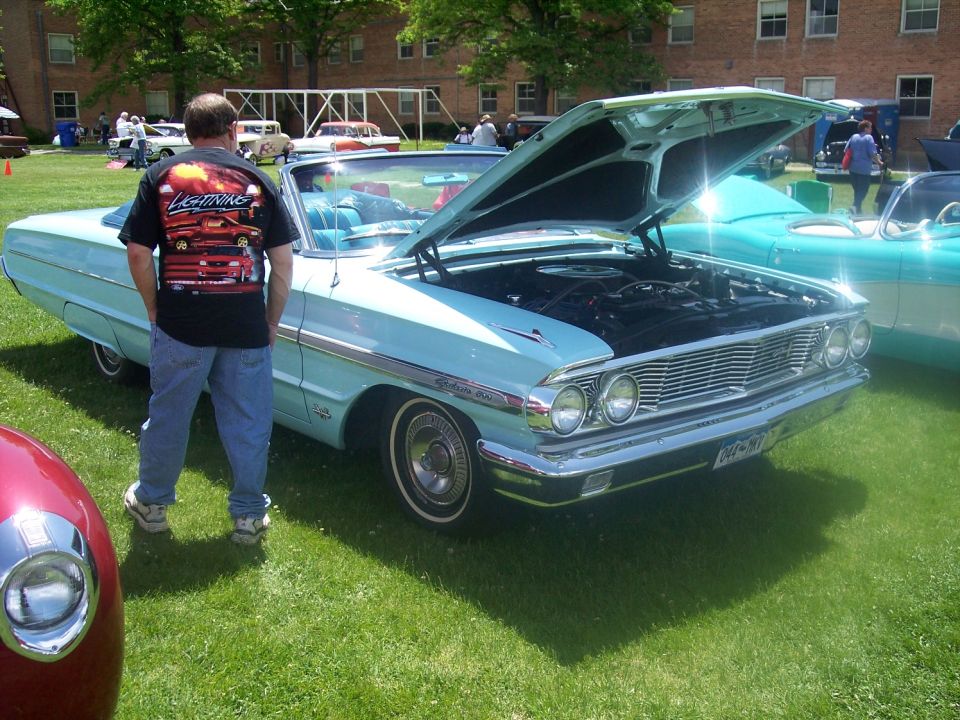 Light blue 1963 Ford Galaxie convertible with hood open, parked on grass. Another car is visible to the right