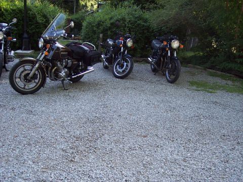 1985 Ford LTD Crown Victoria - A trio of motorcycles are parked on a gravel surface, with lush greenery in the background.