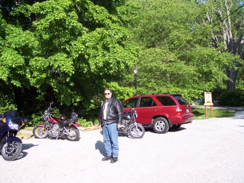 1985 Ford LTD Crown Victoria - A man stands in a gravel lot with several motorcycles and a red SUV under leafy trees.
