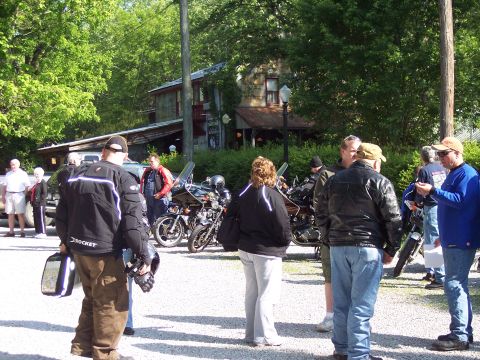 1985 Ford LTD Crown Victoria - Motorcyclists congregate in a lot near a weathered building, surrounded by trees.
