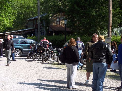 1985 Ford LTD Crown Victoria - A group of people and motorcycles are gathered in a gravel lot near a rustic building.