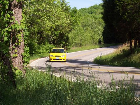 1985 Ford LTD Crown Victoria - A bright yellow sedan navigates a curve on a rural road surrounded by lush green foliage.