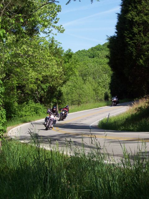 1985 Ford LTD Crown Victoria - Motorcyclists navigate a curve on a scenic road surrounded by lush green trees.