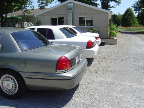 2001 Ford Crown Victoria - Three Ford Crown Victorias, one silver and two white, are parked in a gravel lot next to a building.