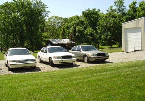 2001 Ford Crown Victoria - Three Ford Crown Victorias are parked in a driveway with a building and trees in the background.
