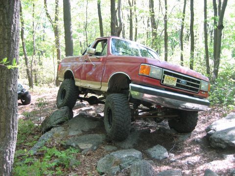 1990 Ford Bronco - Modified pickup truck tackling a challenging off-road course with large rocks and trees.